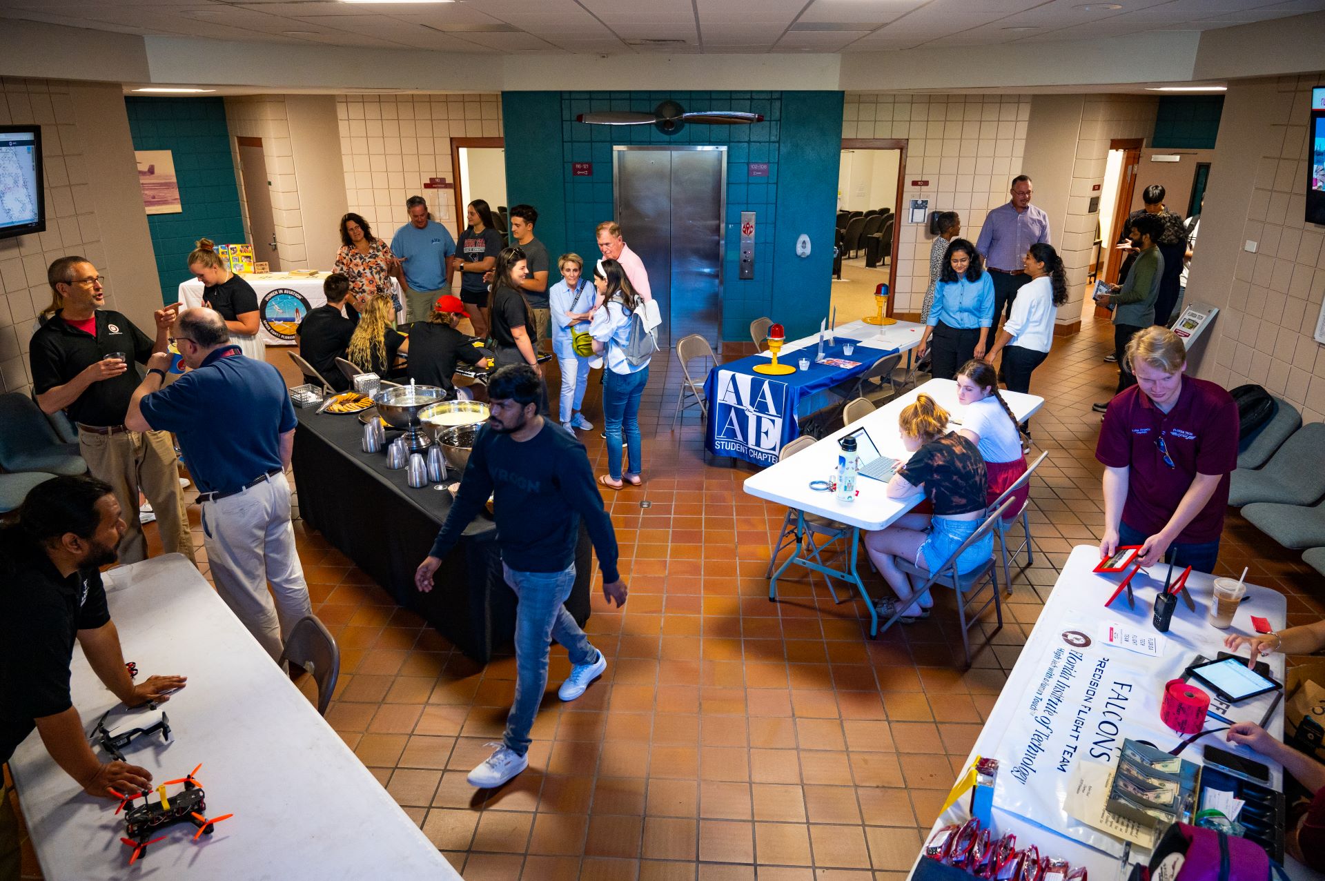 Busy indoor event with students and faculty engaging around tables with various exhibits and projects, including a drone display, in a hallway with food catering set up